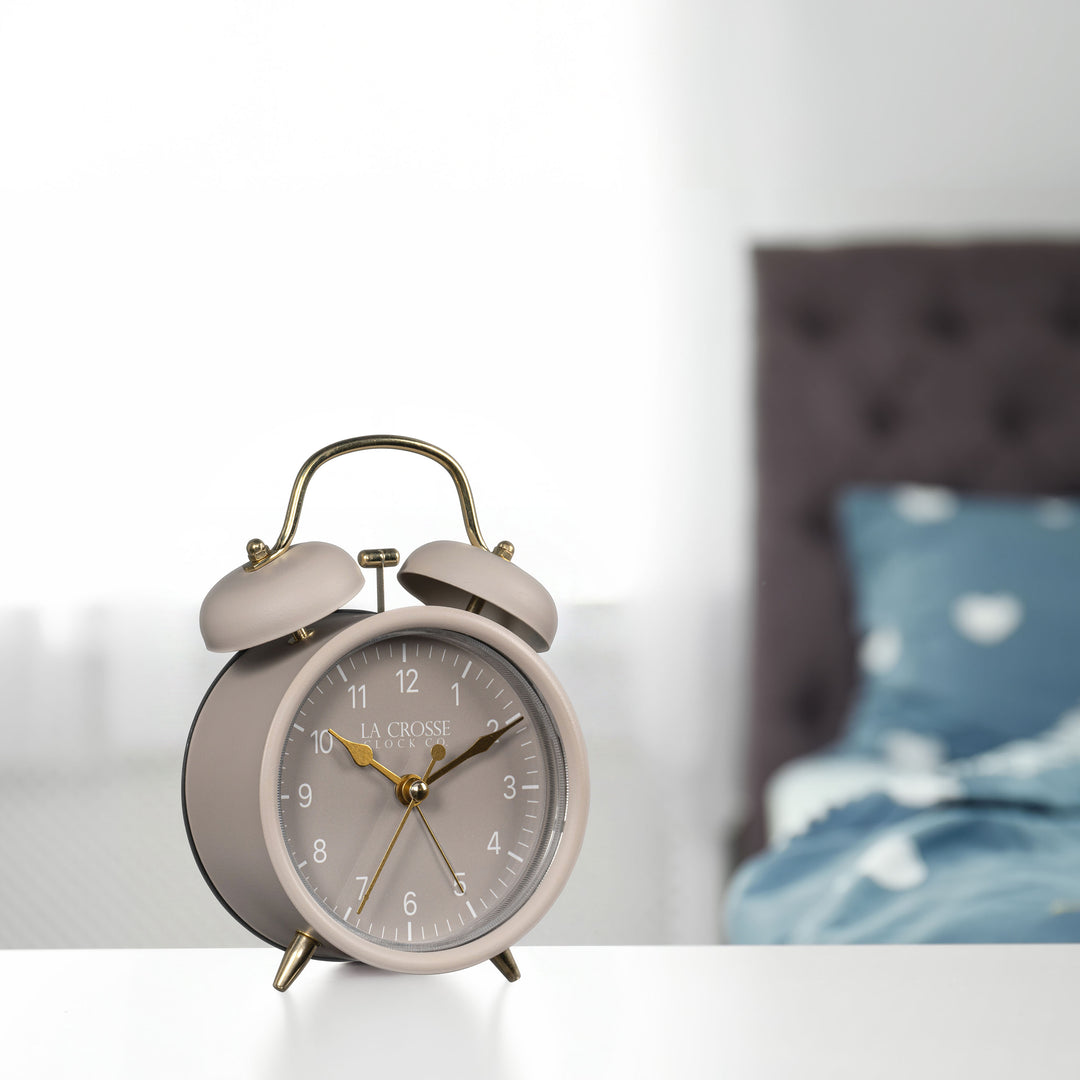 Lifestyle image of the 617-3314TP Metal Twin Bell Alarm Clock – Taupe, sitting on a nightstand with a bedroom scene in the background. The clock features a taupe dial with white numbers, gold hands and accents, in a taupe metal case.