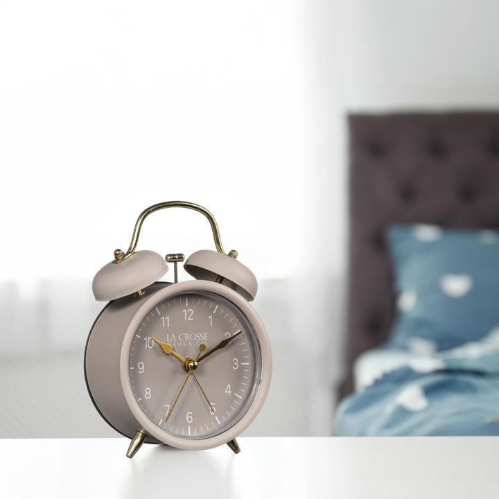 Lifestyle image of the 617-3314TP Metal Twin Bell Alarm Clock – Taupe, sitting on a nightstand with a bedroom scene in the background. The clock features a taupe dial with white numbers, gold hands and accents, in a taupe metal case.