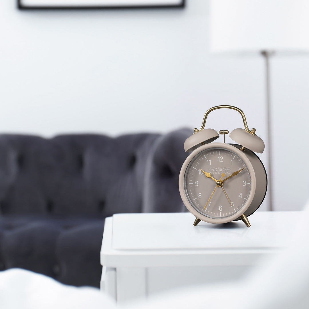 Lifestyle image of the 617-3314TP Metal Twin Bell Alarm Clock – Taupe, sitting on a nightstand with a bedroom scene in the background. The clock features a taupe dial with white numbers, gold hands and accents, in a taupe metal case.