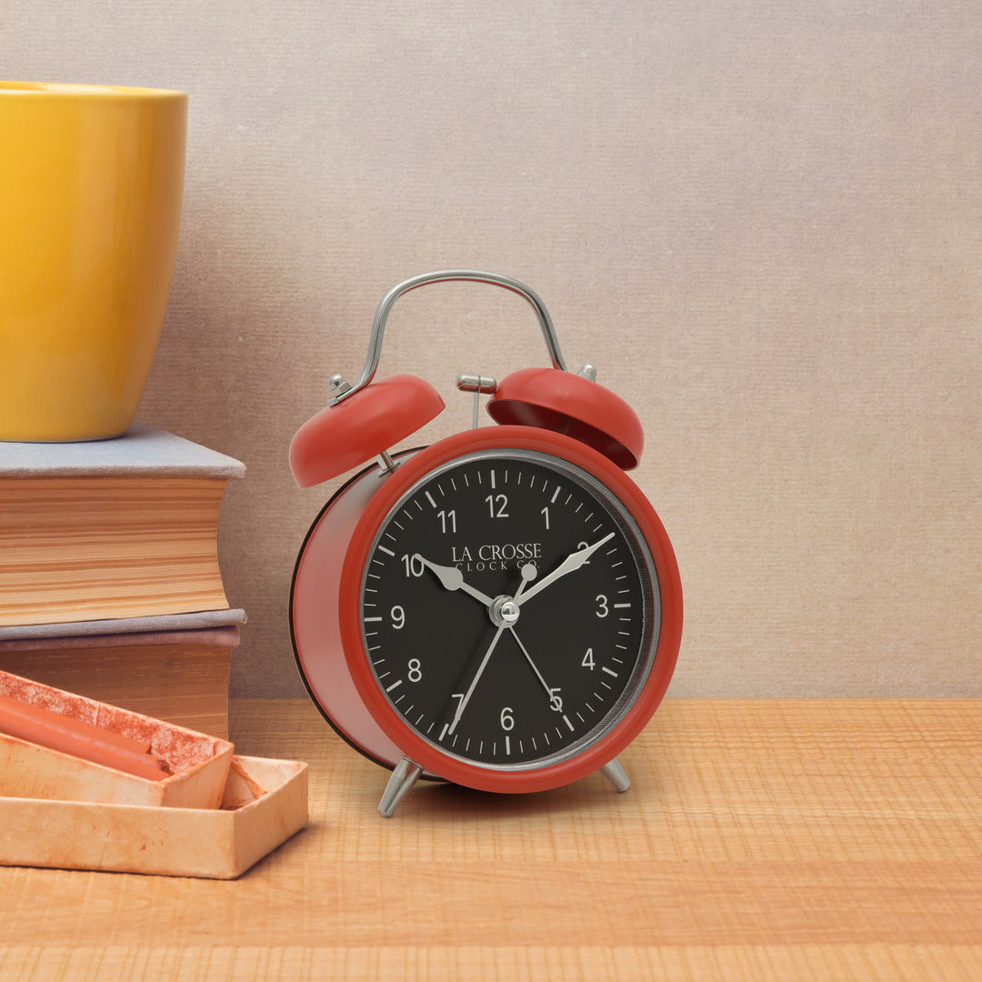 Lifestyle image of the 617-3314 Metal Twin Bell Alarm Clock – Red, sitting on a light wood shelf with books. The clock features a black dial with white numbers, silver hands and accents, in a red metal case
