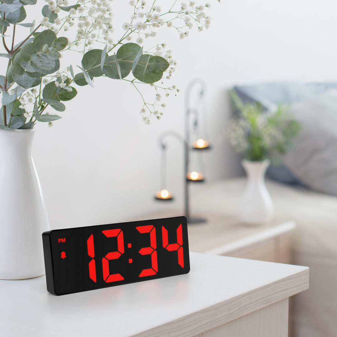 Lifestyle image of the 617-546 2.0-inch Red LED Alarm Clock with indoor temperature shown on a light colored dresser with a nightstand and bed in the background. The clock reads 12:34pm and features 2-inch time digits, with alarm and PM indicators.