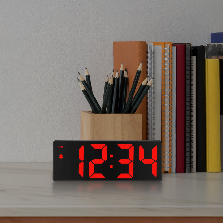 Lifestyle image of the 617-546 2.0-inch Red LED Alarm Clock with indoor temperature shown on a desk with notebooks, books, and a pencil holder. The clock reads 12:34pm and features 2-inch time digits, with alarm and PM indicators.