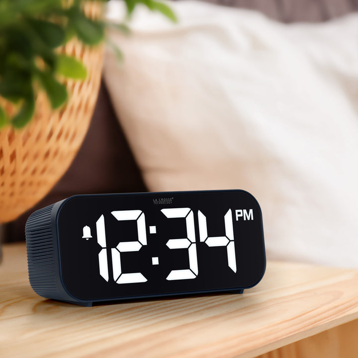Lifestyle image of the 617-714BL Insta-Set Digital LED Alarm Clock with Bluetooth® Technology - Navy sitting on a wood nightstand. The alarm clock features large white digits, a PM indicator, and an alarm icon in a navy blue case.