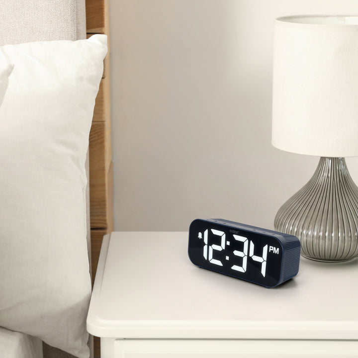 Lifestyle image of the 617-714BL Insta-Set Digital LED Alarm Clock with Bluetooth® Technology - Navy sitting on a white nightstand with a silver base lamp. The alarm clock features large white digits, a PM indicator, and an alarm icon in a navy blue case.