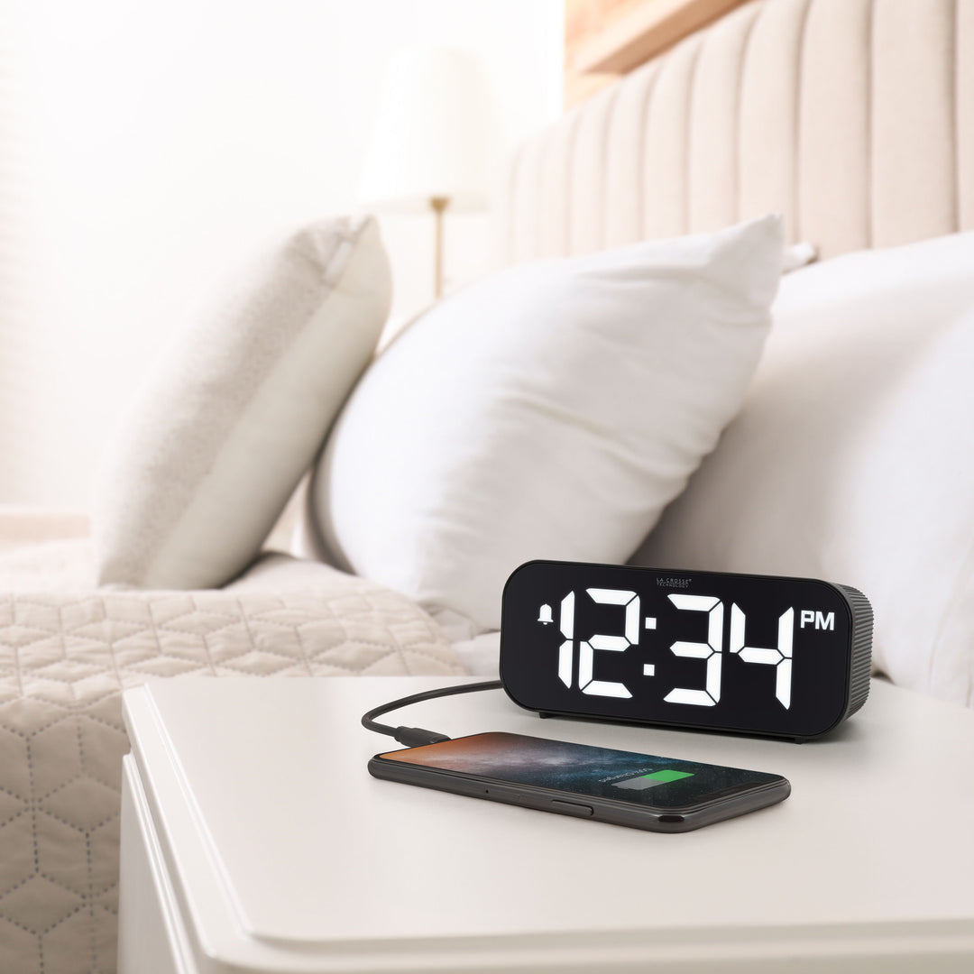 Lifestyle image of the 617-714 Insta-Set Digital LED Alarm Clock with Bluetooth® Technology - Black sitting on a white nightstand with a bed in the background. The alarm clock is shown with a smartphone being charged. The alarm clock features large white digits, a PM indicator, and an alarm icon in a black case.
