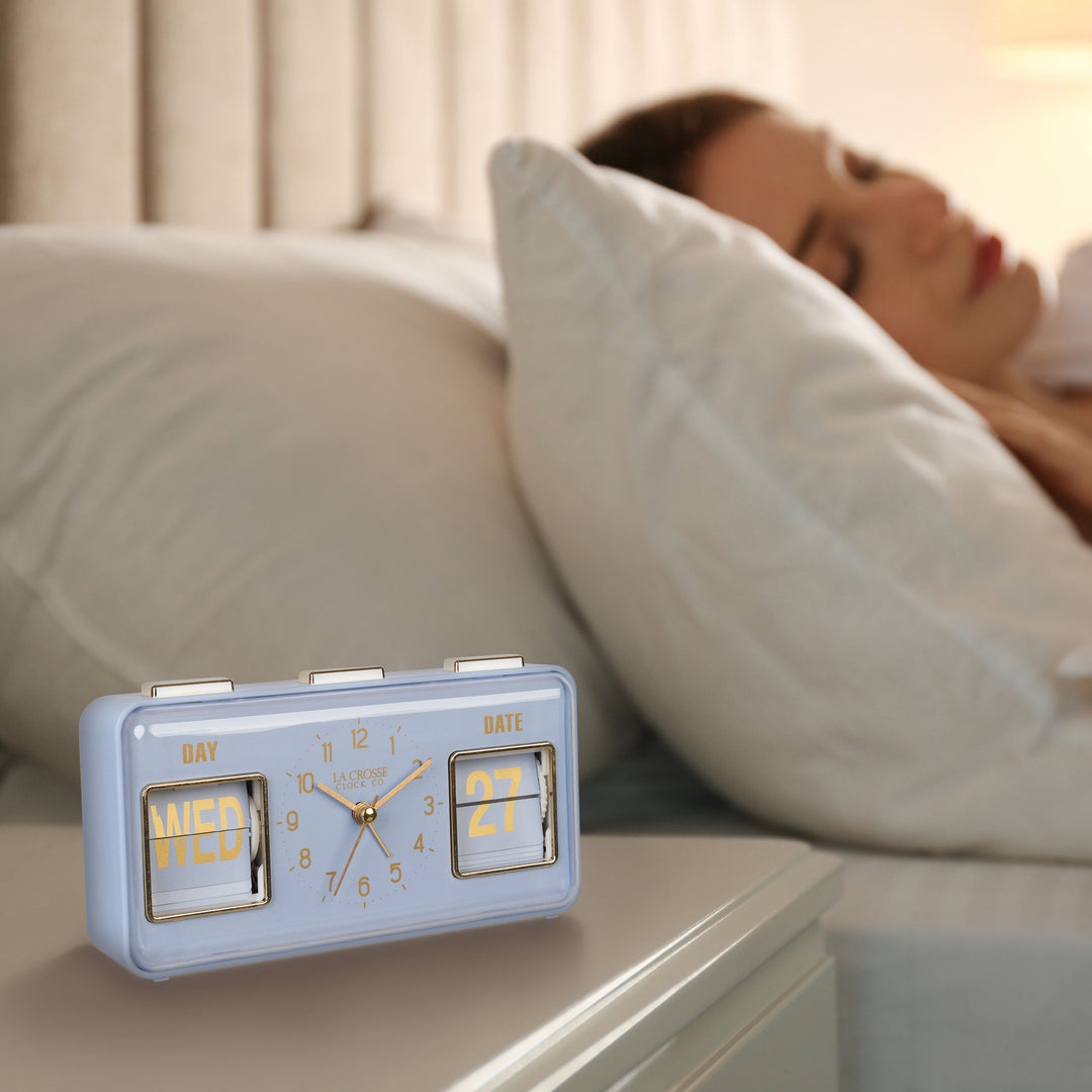 Lifestyle image of the 636-379BL Daily Flip Tabletop Clock with Calendar – Blue, shown sitting on a nightstand with a woman in bed sleeping. The retro rectangle design clock features an analog clock with brass numbers, hour, minute, and second hands, and a day and date calendar with manual flip cards with brass text.