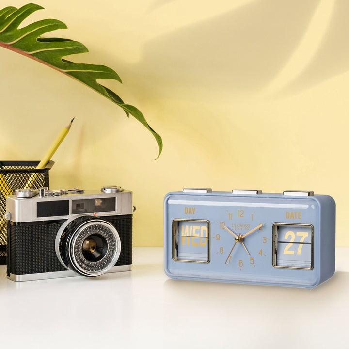 Lifestyle image of the 636-379BL Daily Flip Tabletop Clock with Calendar – Blue, shown sitting on a light-colored desk with a vintage camera and pencil holder in front of a yellow wall. The retro rectangle design clock features an analog clock with brass numbers, hour, minute, and second hands, and a day and date calendar with manual flip cards with brass text.