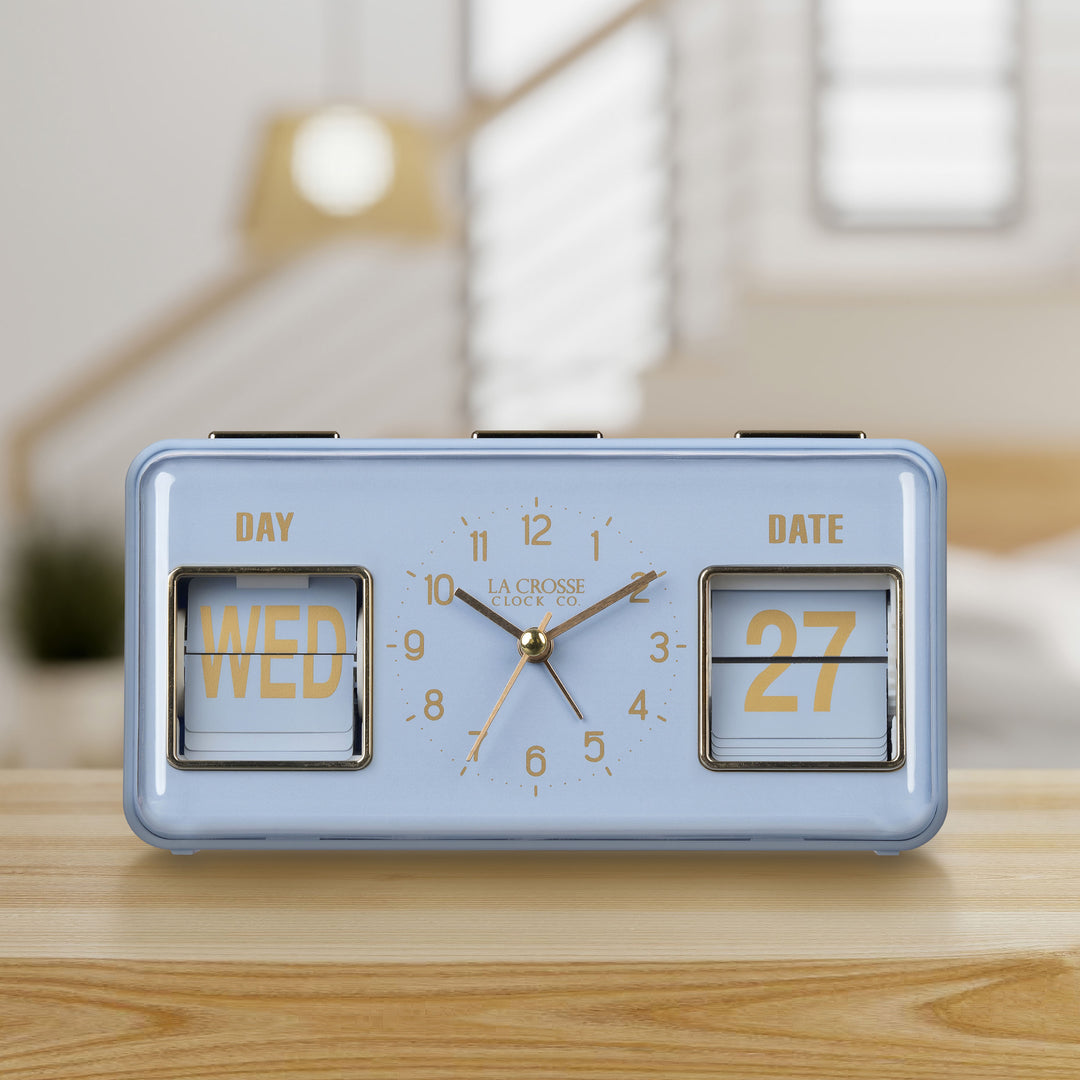 Lifestyle image of the 636-379BL Daily Flip Tabletop Clock with Calendar – Blue, shown sitting on a wood shelf with a bedroom in the background. The retro rectangle design clock features an analog clock with brass numbers, hour, minute, and second hands, and a day and date calendar with manual flip cards with brass text.