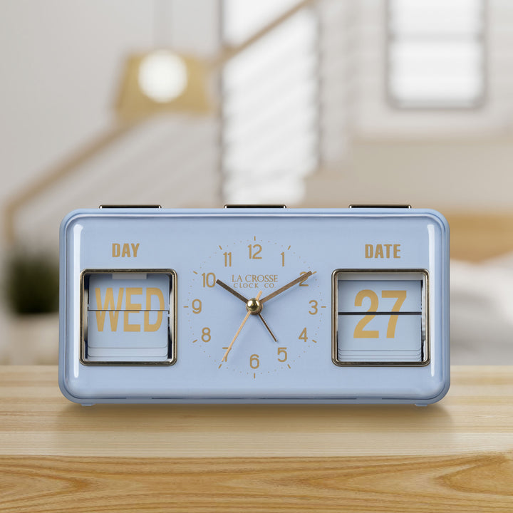Lifestyle image of the 636-379BL Daily Flip Tabletop Clock with Calendar – Blue, shown sitting on a wood shelf with a bedroom in the background. The retro rectangle design clock features an analog clock with brass numbers, hour, minute, and second hands, and a day and date calendar with manual flip cards with brass text.