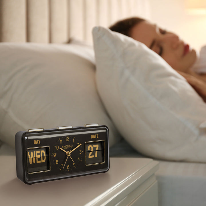 Lifestyle image of the 636-379B Daily Flip Tabletop Clock with Calendar – Black, shown sitting on a nightstand with a woman in bed sleeping. The retro rectangle design clock features an analog clock with brass numbers, hour, minute, and second hands, and a day and date calendar with manual flip cards with brass text.