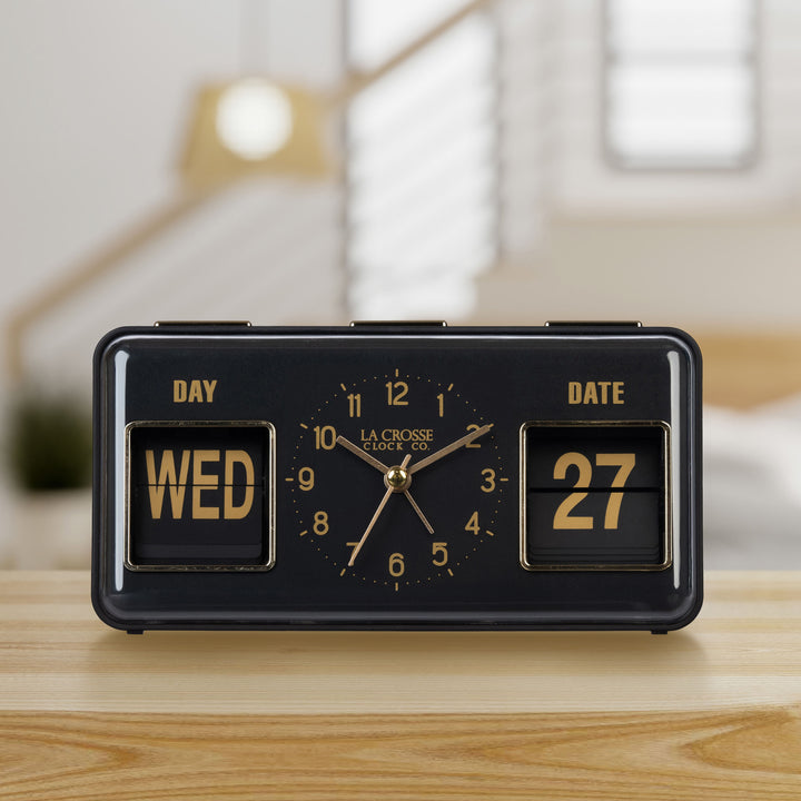 Lifestyle image of the 636-379B Daily Flip Tabletop Clock with Calendar – Black, shown sitting on a wood shelf with a bedroom in the background. The retro rectangle design clock features an analog clock with brass numbers, hour, minute, and second hands, and a day and date calendar with manual flip cards with brass text.