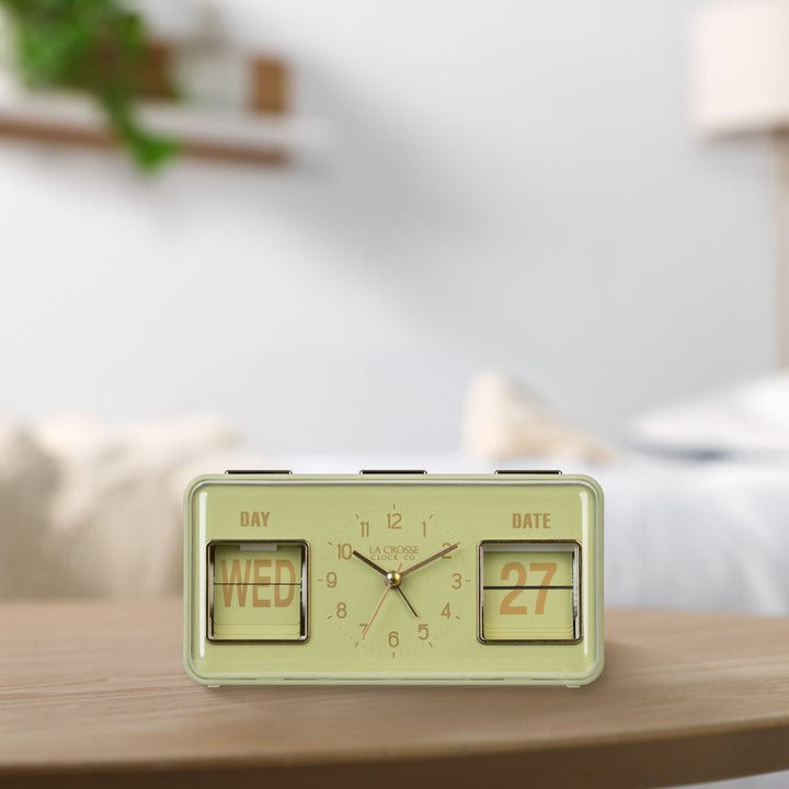 Lifestyle image of the 636-379G Daily Flip Tabletop Clock with Calendar – Green shown sitting on a light wood table. The retro rectangle design clock features an analog clock with brass numbers, hour, minute, and second hands, and a day and date calendar with manual flip cards with brass text.