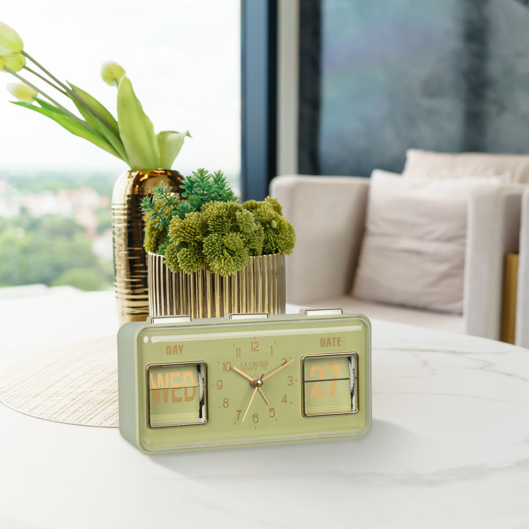 Lifestyle image of the 636-379G Daily Flip Tabletop Clock with Calendar – Green shown sitting on a faux marble coffee table in front of brass planters. The retro rectangle design clock features an analog clock with brass numbers, hour, minute, and second hands, and a day and date calendar with manual flip cards with brass text.