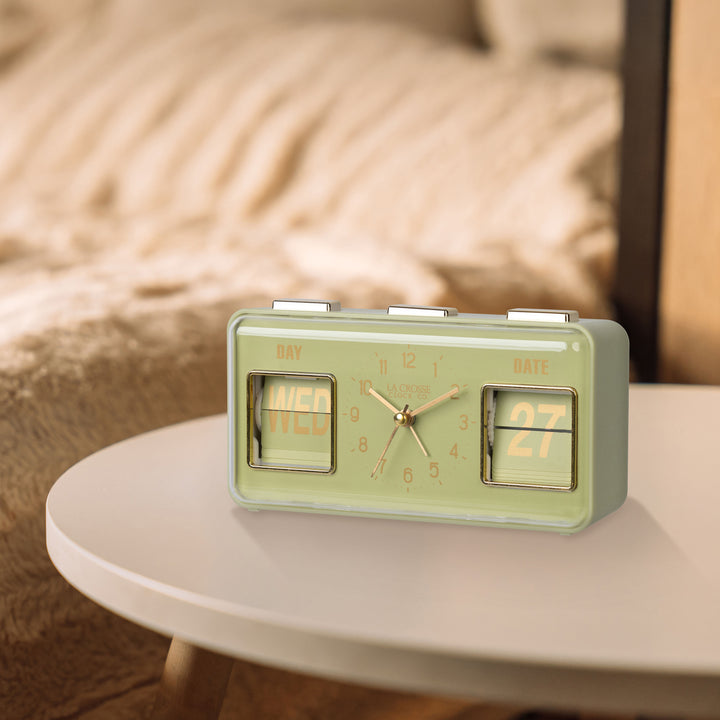 Lifestyle image of the 636-379G Daily Flip Tabletop Clock with Calendar – Green shown sitting on a neutral side table with faux fur tan bedding in the background. The retro rectangle design clock features an analog clock with brass numbers, hour, minute, and second hands, and a day and date calendar with manual flip cards with brass text.