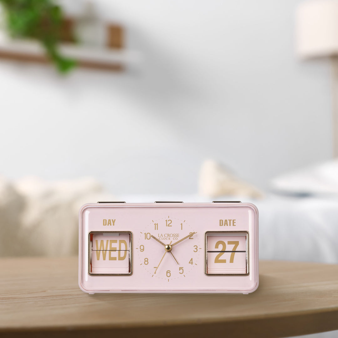 Lifestyle image of the 636-379P Daily Flip Tabletop Clock with Calendar – Pink Blush, shown sitting on a light wood table. The retro rectangle design clock features an analog clock with brass numbers, hour, minute, and second hands, and a day and date calendar with manual flip cards with brass text.