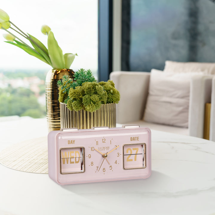 Lifestyle image of the 636-379P Daily Flip Tabletop Clock with Calendar – Pink Blush, shown sitting on a faux marble coffee table in front of brass planters. The retro rectangle design clock features an analog clock with brass numbers, hour, minute, and second hands, and a day and date calendar with manual flip cards with brass text.