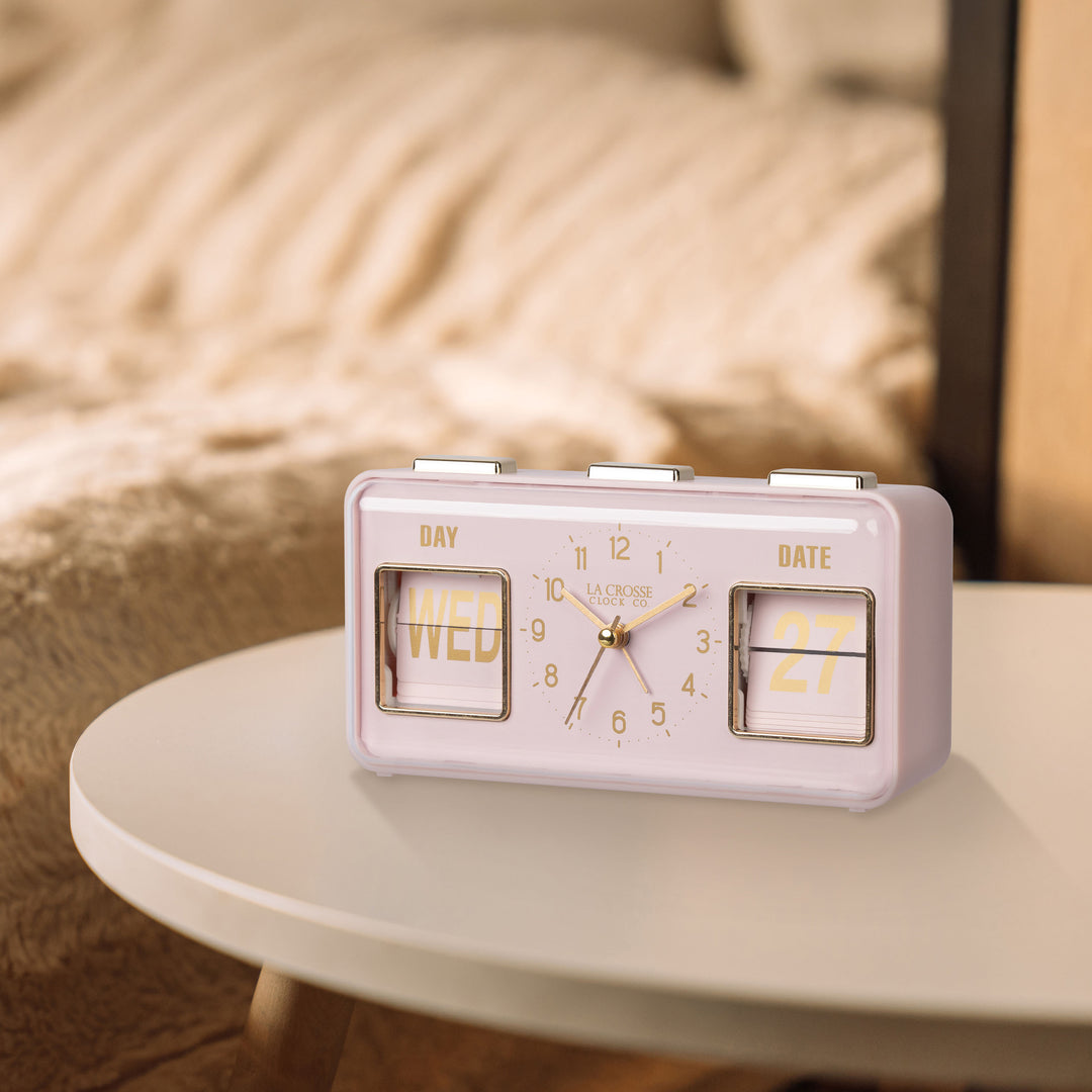 Lifestyle image of the 636-379P Daily Flip Tabletop Clock with Calendar – Pink Blush, shown sitting on a neutral side table with faux fur tan bedding in the background. The retro rectangle design clock features an analog clock with brass numbers, hour, minute, and second hands, and a day and date calendar with manual flip cards with brass text.
