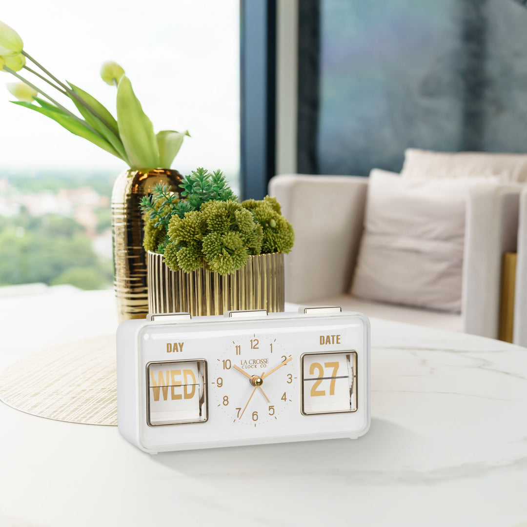 Lifestyle image of the 636-379W Daily Flip Tabletop Clock with Calendar – White, shown sitting on a faux marble coffee table in front of brass planters. The retro rectangle design clock features an analog clock with brass numbers, hour, minute, and second hands, and a day and date calendar with manual flip cards with brass text.