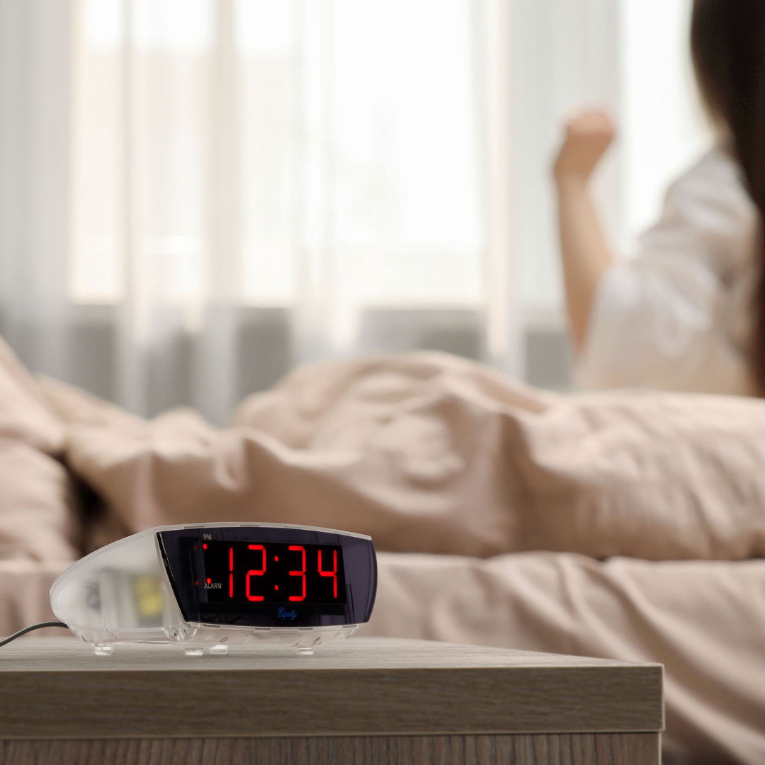 Lifestyle image of the 75908 0.9-inch Red LED Clear Case Alarm Clock shown on a night table next to a bed. The digital alarm clock features red digits with a black lens, PM and ALARM indicators, and is in a clear case.