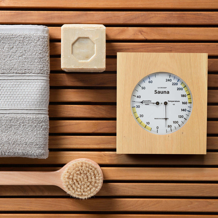 Lifestyle image of the 914-620 Sauna Thermometer and Hygrometer dial in oiled oak wood casing displaying a dial split in half with temperature on the right in Fahrenheit from 60 degrees to 245 degrees with optimal range in yellow and the left half showing humidity readings from 0 percent to 75 percent with the optimal range highlighted in yellow sitting on a sauna bench with a towel brush and soap