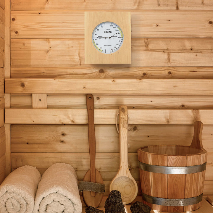 Lifestyle image inside a sauna of the 914-620 Sauna Thermometer and Hygrometer dial in oiled oak wood casing displaying a dial split in half with temperature on the right in Fahrenheit from 60 degrees to 245 degrees with optimal range in yellow and the left half showing humidity readings from 0 percent to 75 percent with the optimal range highlighted in yellow hanging on the wall above a bench with towels, brush, bucket, and other suana tools