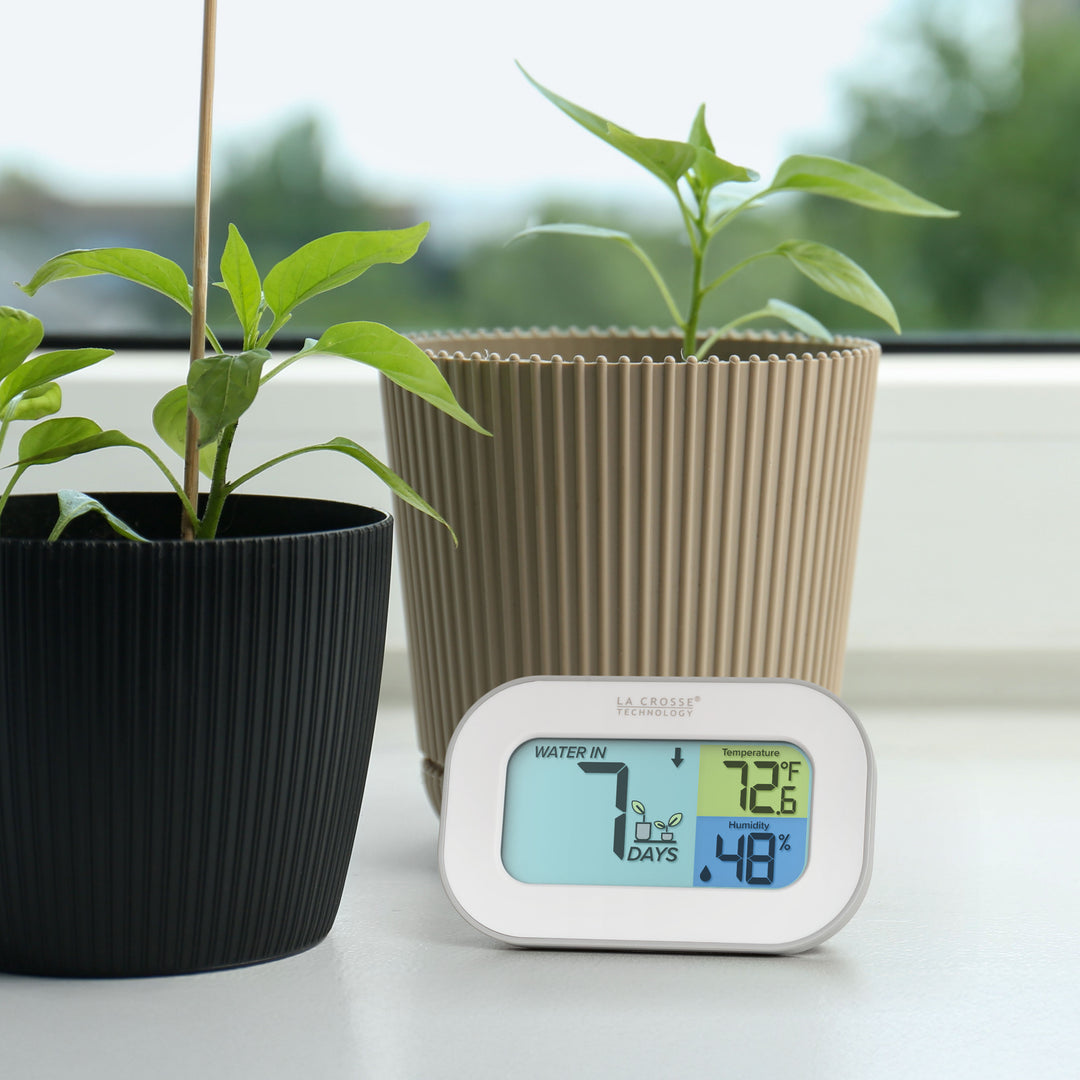 Image of the 919-147 Digital plant care timer with temperature and humidity. The timer sits on a windowsill with potted plants. Battery-operated, the timer features a white and gray case with a three section screen. The turquoise blue section displays how many days since the last watering (plant graphic with an up arrow) the yellow section lists the temperature at 72.6° F, and the dark blue section lists the humidity level at 48 percent with a water drop icon.