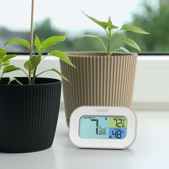 Image of the 919-147 Digital plant care timer with temperature and humidity. The timer sits on a windowsill with potted plants. Battery-operated, the timer features a white and gray case with a three section screen. The turquoise blue section displays how many days since the last watering (plant graphic with an up arrow) the yellow section lists the temperature at 72.6° F, and the dark blue section lists the humidity level at 48 percent with a water drop icon.