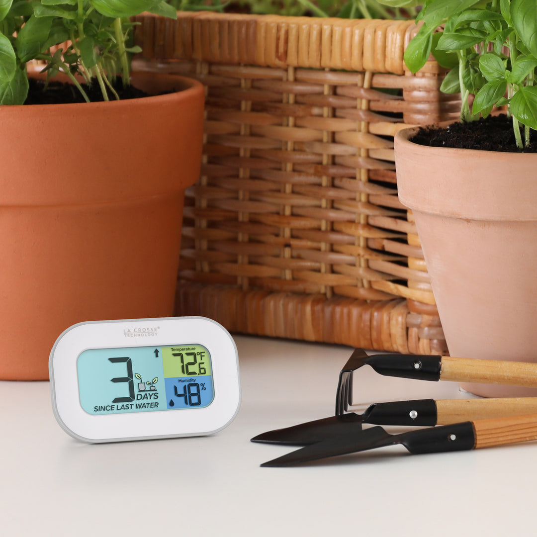 Lifestyle of the 919-147 Digital plant care timer with temperature and humidity. The timer sits on a table with terracotta pots, a basket, and planting tools. Battery-operated, the timer features a white and gray case with a three section screen. The turquoise blue section displays how many days since the last watering (plant graphic with an up arrow) the yellow section lists the temperature at 72.6° F, and the dark blue section lists the humidity level at 48 percent with a water drop icon.