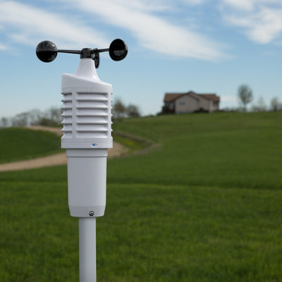 Lifestyle image of a wind speed sensor in the foreground mounted on a white pole with a house in the background with green grass and a long gravel driveway