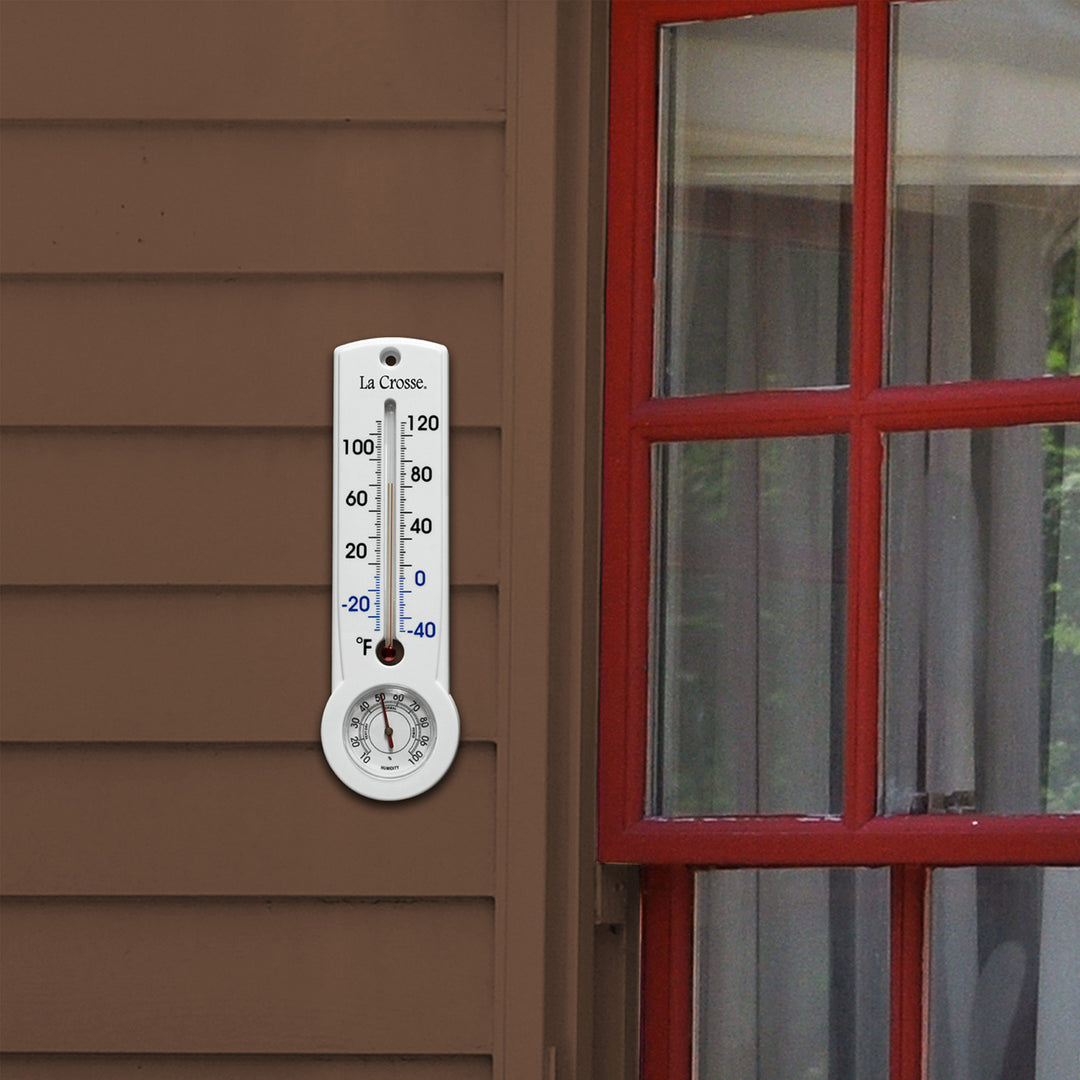 Lifestyle image of the 204-109 8.75-inch Thermometer with Hygrometer and Key Hider in a white case hanging on the siding of a brown house with red window trim. The mercury-free capillary thermometer has a Fahrenheit temperature scale. Temperature markings below freezing appear in blue text, while those above freezing appear in black text. The Hygrometer dial reads 10-100 percent humidity, with a red pointer.