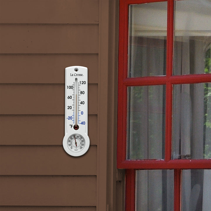 Lifestyle image of the 204-109 8.75-inch Thermometer with Hygrometer and Key Hider in a white case hanging on the siding of a brown house with red window trim. The mercury-free capillary thermometer has a Fahrenheit temperature scale. Temperature markings below freezing appear in blue text, while those above freezing appear in black text. The Hygrometer dial reads 10-100 percent humidity, with a red pointer.