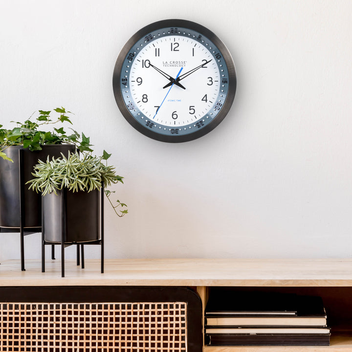 Lifestyle image of the 404-54667 10-inch Atomic Metal Wall Clock with Blue Chapter Ring hanging on a wall over a light wood side table with plants. The clock features a black stainless steel frame with a blue chapter ring and raised seconds indicators, surrounding a white dial with black numbers, metal hands, and a blue accent second hand.