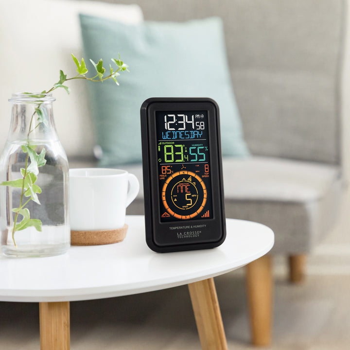 Lifestyle image of the S81120 Color Weather Station with Breeze shown sitting on a white side table next to a gray couch. The color display features a vertical design with time, date, outdoor temperature, humidity, feels like reading, wind speed, and wind direction